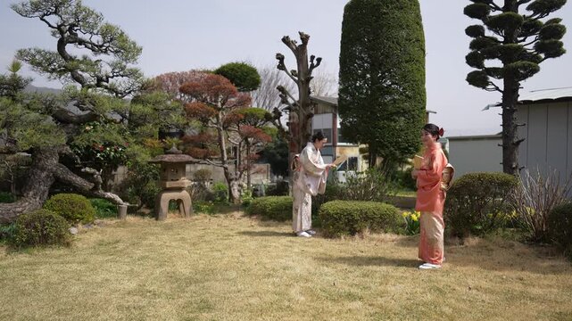 Two women in kimonos stand holding wooden paddles on a lawn surrounded by pruned trees and shrubs. Japan, April 19, 2025, spring.