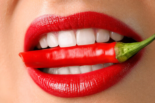 Extreme close-up macro of woman's mouth with glossy red lipstick biting a vibrant red hot chili pepper. Conceptual shot symbolizing passion, spice, heat, and temptation.