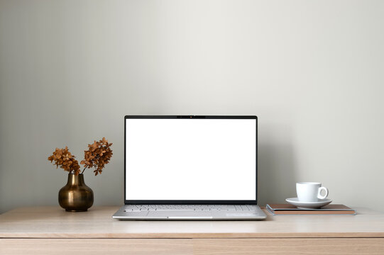 Open laptop with blank screen mockup, coffee, vase with flowers on wooden table, neutral beige wall background with soft natural light, minimal aesthetic home office workspace