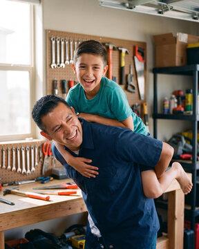 Joyful father giving son piggyback ride in home garage workshop with warm natural light