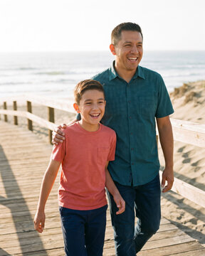 Devoted father and son walking together along sandy beach boardwalk with warm natural light