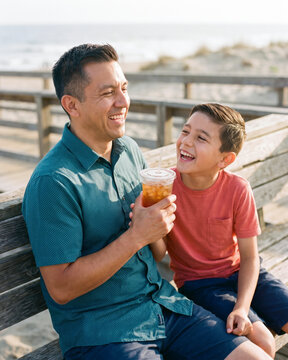 Playful father and son sharing iced tea on sunny beach boardwalk with warm natural light