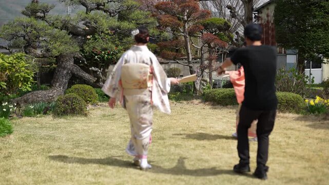 A man in dark casual clothing crosses the foreground while women in kimonos hold wooden paddles on a garden lawn. Japan, April 19, 2025, spring.
