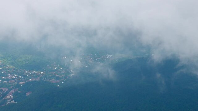 Stunning aerial views of clouds and mist flowing overgreen mountain valley, with small town tucked away among hills, sunlight breaking through fog, creating spectacular landscape in Georgia Tkibuli's