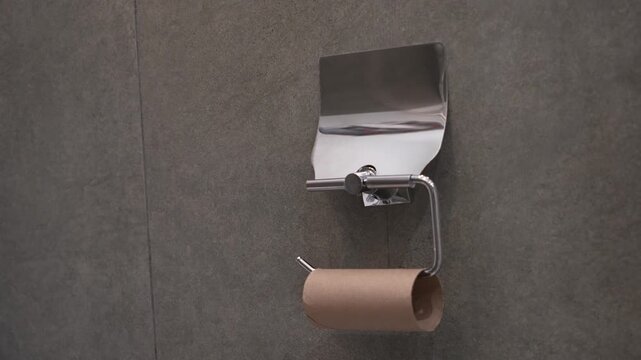 Empty toilet paper roll holder close up, brown cardboard core tube on chrome metal hanger inside bathroom restroom, running out of household tissue supply conceptual minimal apartment background.
