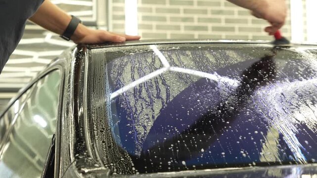 Professional auto detailer hands preparing a wet car windshield glass for an anti-scratch and chip protection film application. Male technician washing a window surface with a brush and soapy water. 