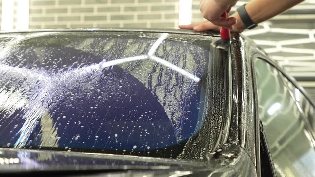 Professional auto detailer hands preparing a wet car windshield glass for an anti-scratch and chip protection film application. Male technician washing a window surface with a brush and soapy water. 