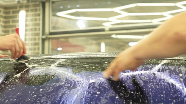 Professional auto detailer hands preparing a wet car windshield glass for an anti-scratch and chip protection film application. Male technician washing a window surface with a brush and soapy water. 