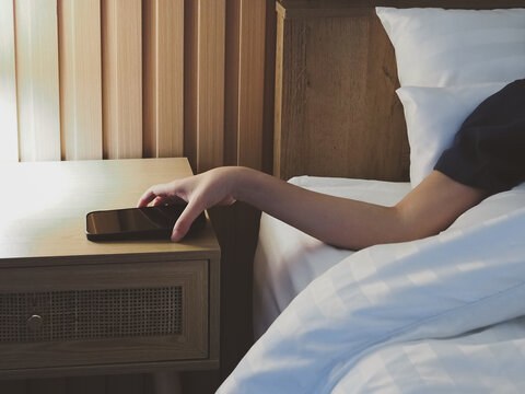 A young woman's hand reaches for her smartphone on the bedside table in a modern bedroom decorated with warm wooden tones and bathed in soft morning sunlight, creating a peaceful atmosphere.
