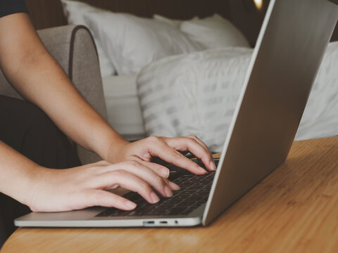 Person typing on laptop keyboard on wooden table in cozy bedroom setting, focused hands working on computer with soft bed background and warm natural light