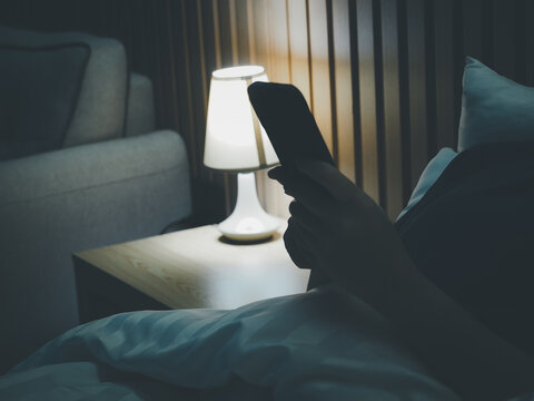 Person holding smartphone bedside lamp bedroom night modern wooden headboard cozy bedding dim lighting relaxation late night technology