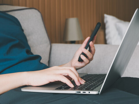 A woman is typing on her laptop and holding her smartphone, relaxing on a sofa in her home under the soft lighting.