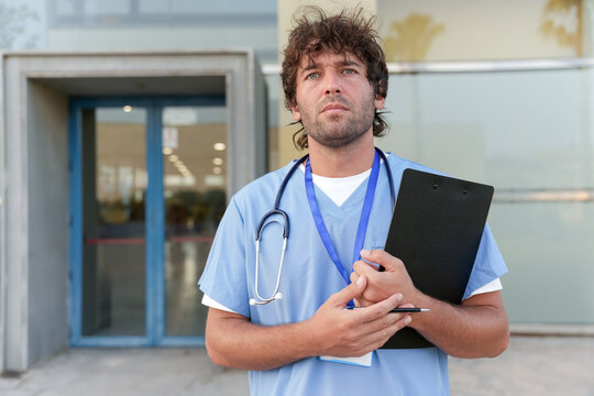 Male nurse wearing medical scrubs and stethoscope, holding a clipboard outside a hospital building