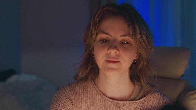 Young adult woman seated at desk in dim blue-lit workspace, looking toward monitor as expression changes from squinting tension to calm concentration, with subtle head tilt and steady focus