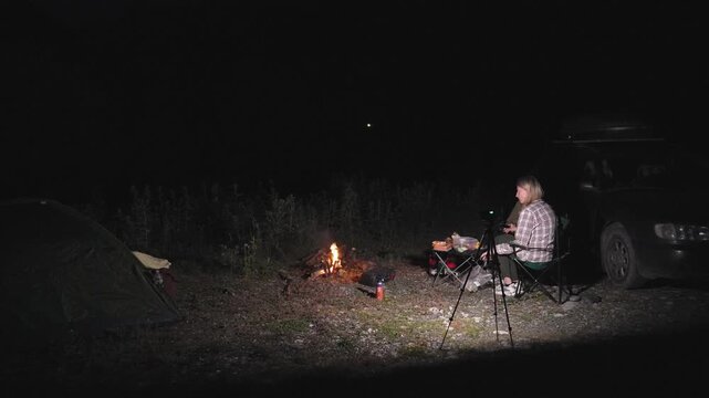 Blonde woman wearing a plaid shirt sitting in a camping chair, filming a crackling bonfire with a camera on a tripod during a night car camping trip while preparing food on a small table