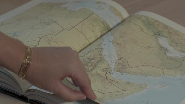 a woman's hand with a gold bracelet is flipping through the pages of a large atlas or world map book on the table. 