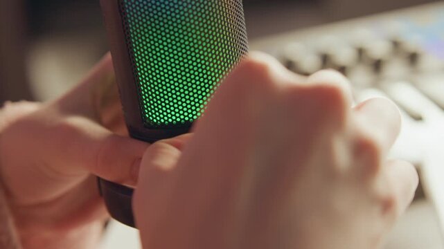 Close-up hands tighten and position condenser microphone inside shock mount while green illumination glows across grille, creating active studio scene with warm soft lighting and shallow depth of