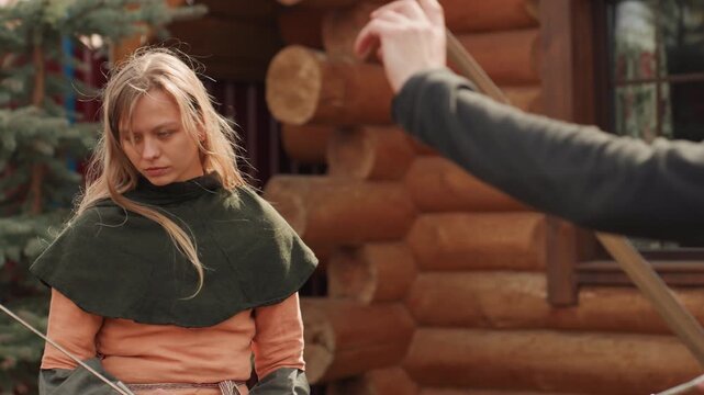 Young woman holds spear with both hands, turning slightly with alert expression beside rustic log cabin; layered cloak and tunic, loose hair, warm daylight, outdoor tension, readiness, courage