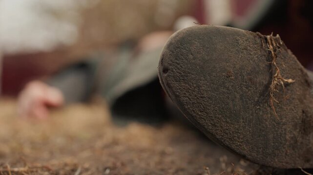 Low-angle close view of mud-covered boot sole resting on dry grass and soil beside prone person in blurred background, with rough texture, earthy tones, and tense outdoor scene, with boot, sole, mud