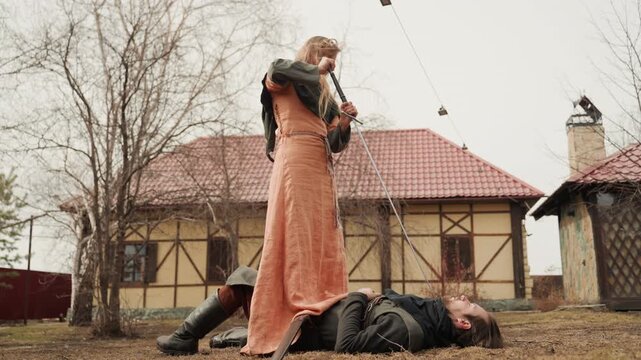 Woman in medieval dress stands over prone man with sword lifted high, creating tense outdoor confrontation; raised arm, angled blade, and rigid stance suggest imminent attack, with woman, sword