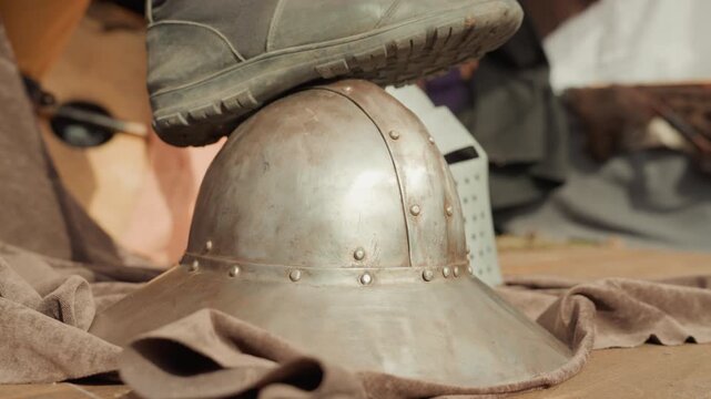 Boot presses worn metal helmet on folded brown fabric, tilting brim and compressing cloth while scuffed dome shows rivets, dents, rust, and weathered texture under warm directional light
