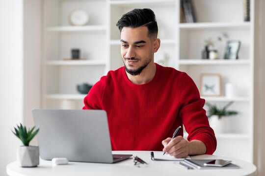 Focused Young Man Of Middle Eastern Ethnicity Working On Laptop While Taking Notes At Home Office, Showing Professionalism In His Creative Workspace. Emphasizing Business Concept.