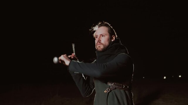 Man wearing harness and jacket works outdoors at night, pulling and adjusting rope while turning toward bright backlight that creates rim glow and soft flare; medium three quarter viewpoint with