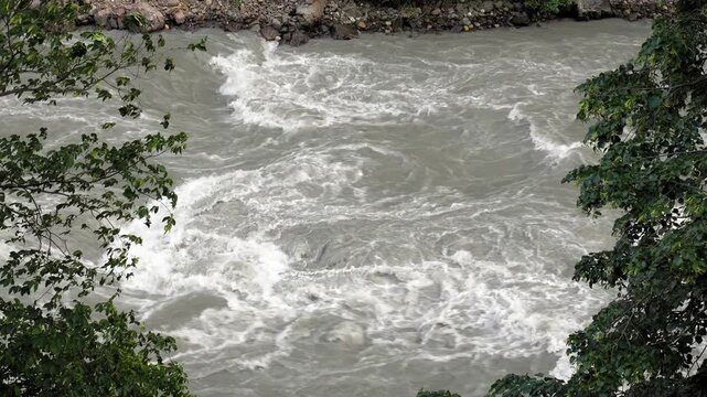 Top-down view of a powerful, wild mountain river with turbulent rapids and gray, muddy water flowing quickly through a rocky channel, framed by green foliage on the riverbanks