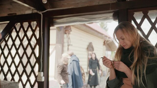Blonde young woman standing inside wooden pavilion inspects small dangling keychain with both hands while smiling, leaning slightly forward, with gentle hand rotation and subtle head tilt in diffuse