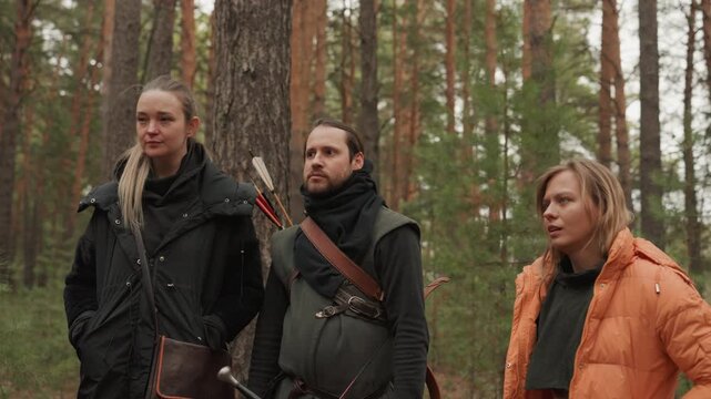 Three young adults stand in pine forest clearing, facing forward with alert expressions while scanning surroundings; central figure carries leather quiver and arrows, ready for action