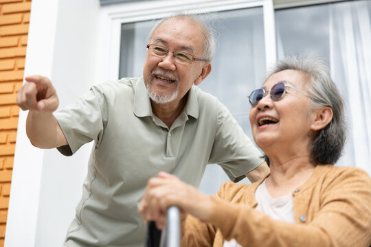 Senior Asian couple laughing outdoors near home with walking aid, sharing joyful moment and pointing together during active retirement lifestyle
