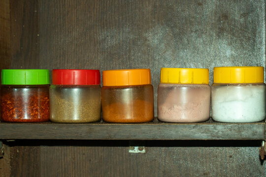 Row of five colorful plastic spice jars on dark wooden shelf close.