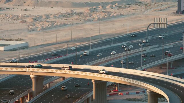 Cars and vehicles travel along a curved elevated highway overpass and multi lane road crossing desert terrain during sunny day in Dubai, UAE