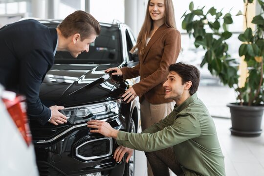 Smiling Caucasian Couple Engaging With Male Salesperson Discussing Features Of A Black Suv At Auto Dealership. They Are Focused And Interested In Buying A New Car In A Professional Setting.