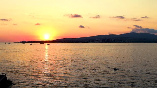 Sunset over calm water with boats sailing, silhouetted trees framing the scene, and distant hills creating a serene landscape in a coastal region