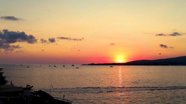 Sunset over calm water with boats sailing, silhouetted trees framing the scene, and distant hills creating a serene landscape in a coastal region