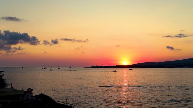 Sunset over calm water with boats sailing, silhouetted trees framing the scene, and distant hills creating a serene landscape in a coastal region