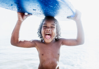 Fototapeta premium Joyful Black boy holding a blue surfboard overhead at the beach