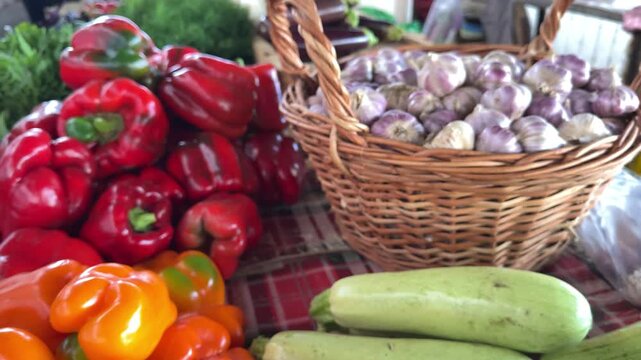 A colorful market stall displays ripe bell peppers, fresh garlic bulbs in a wicker basket and green zucchini, showing an abundant selection of seasonal farm produce. Close up.