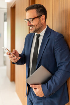 Professional man standing in an office corridor, smiling while using his mobile phone and holding a laptop, engaging in corporate communication and business networking