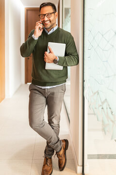Smiling professional standing in an office corridor, holding a laptop, engaging in a phone conversation, representing modern business communication and success