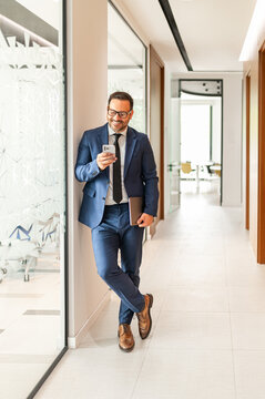 Business professional smiling and using a smartphone while standing in a modern corporate corridor, holding a portfolio, engaging in mobile communication and work networking