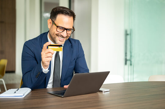 Smiling businessman making online purchase with credit card, using laptop for secure banking, handling finances, and managing accounts in a modern office environment