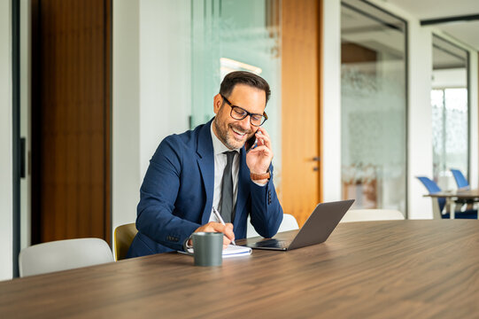 Professional male working in modern corporate office. Laughing during a phone conversation. Multitasking while taking notes and using a laptop