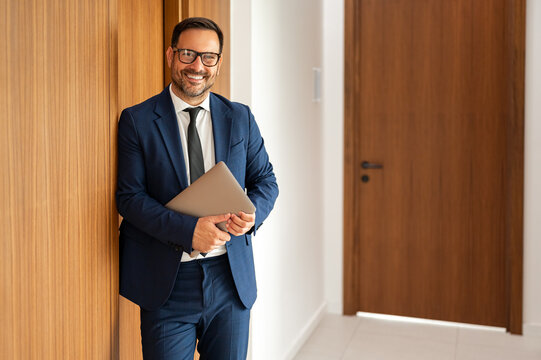 Business professional smiling confidently, holding a laptop, standing in a contemporary office corridor, representing success and executive leadership