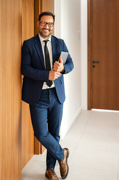 Smiling man holding a digital tablet in a blue suit and tie, standing confidently in an office corridor, representing modern business communication and success