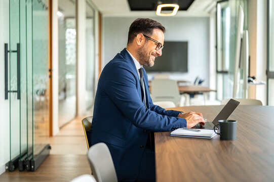 Experienced male entrepreneur managing business operations, smiling while typing on laptop during important financial planning, working in a modern corporate office environment