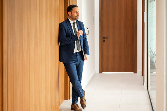 Male professional in a business suit holding eyeglasses and a portfolio, contemplating his financial strategy and legal responsibilities in an office environment