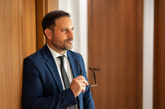 Professional businessman in suit holding eyeglasses, gazing thoughtfully out of an office window, reflecting on strategy, leadership and important career decisions