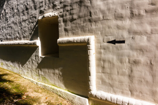 Fragment of a white stone fortress wall of an ancient monastery with a small window embrasure and a black directional arrow sign pointing left along the textured masonry surface